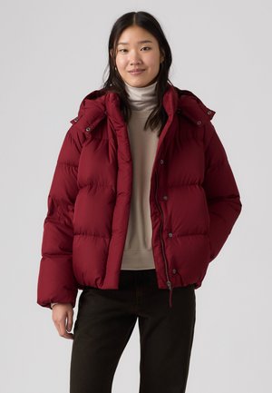 Giovane donna che indossa una giacca imbottita rossa sopra un maglione beige con collo alto e pantaloni neri, in piedi contro uno sfondo grigio chiaro uniforme.