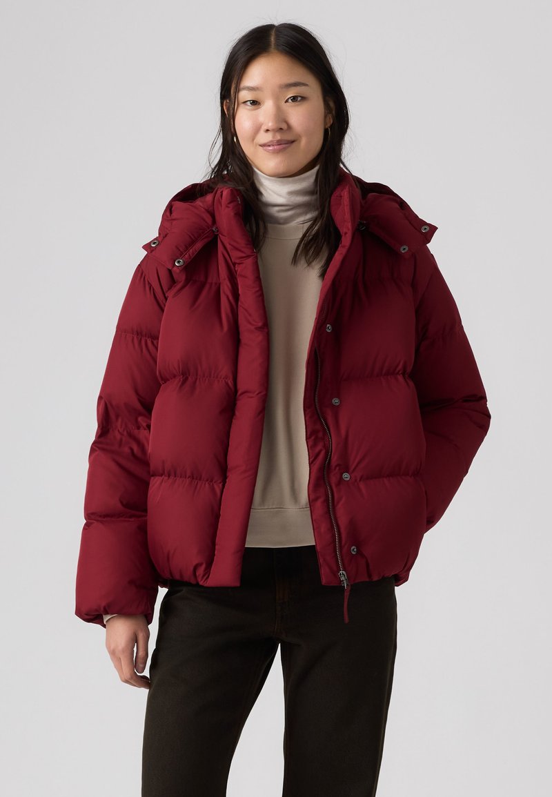 Jeune femme portant une doudoune rouge par-dessus un pull à col roulé beige et un pantalon noir, debout devant un fond gris clair uni.
