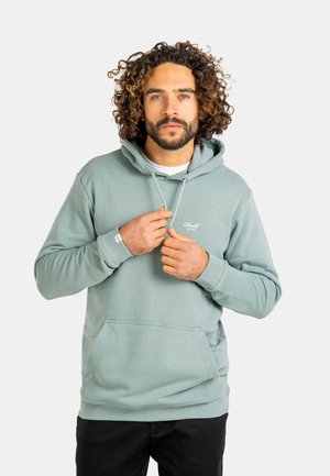 Man with curly hair wearing a light green hoodie, adjusting its drawstrings, standing against a plain white background.