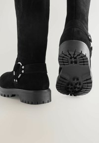 Zwarte suède kniehoge laarzen hebben een gespsluiting en een stevige rubberen zool met diepe profielen voor grip. De hak is plat.