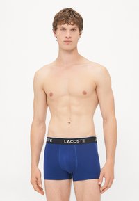 Mornarško modri bokserice z črnim pasom, ki nosi logotip Lacoste. Pameten material, prilegajoča se zasnova, gladka tekstura, dolžina do sredine stegna.
