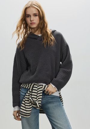 Grå strikket hættetrøje med løs pasform og v-hals. Kombineret med blå jeans, en stribet skjorte bundet ved taljen og metalliske ringe.