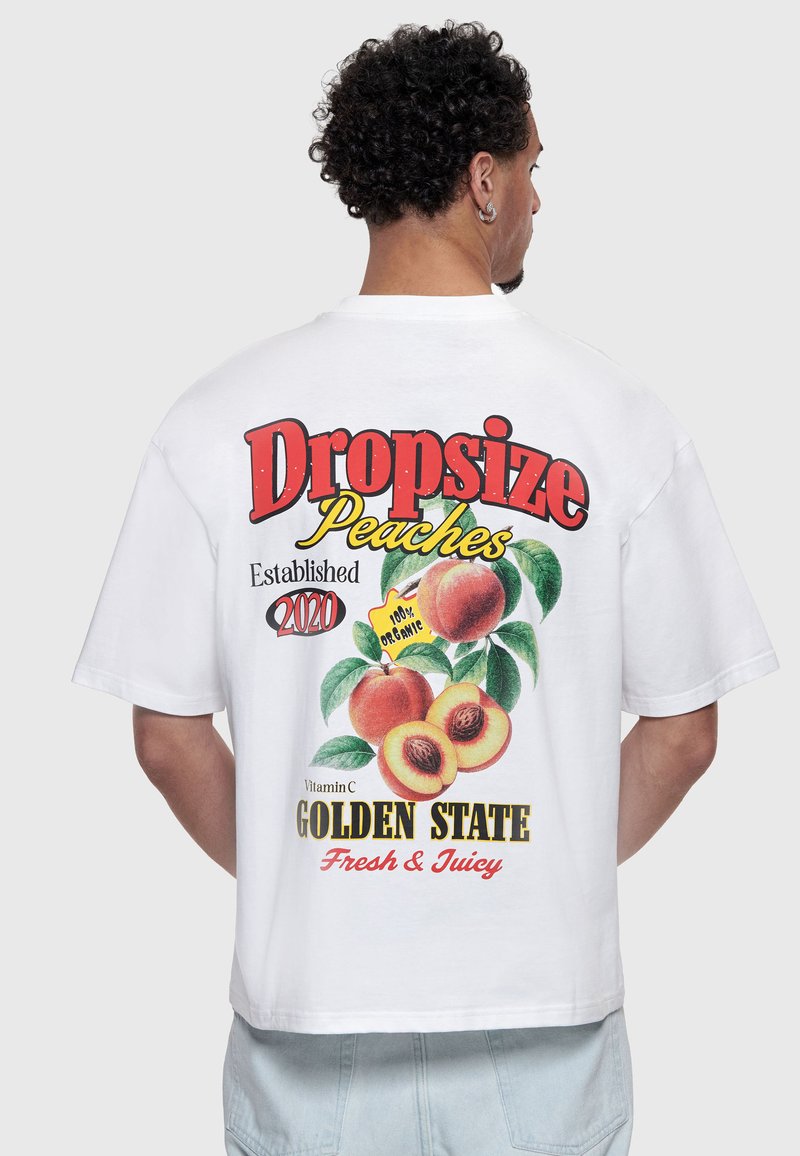 Homme aux cheveux bouclés portant un t-shirt blanc avec des graphismes colorés de pêches et un texte sur les pêches biologiques dans le dos.