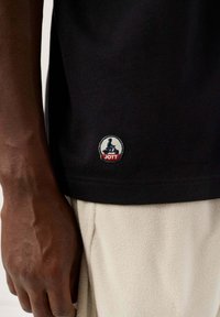 Schwarzes Baumwoll-T-Shirt mit einem runden JOTT-Logo in Blau und Rot, nahe dem Saum platziert, zusammen mit weißen Fleece-Hosen.