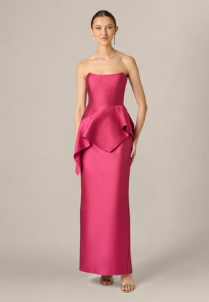 Vrouw draagt een strapless fuchsia jurk met asymmetrisch peplumdetail en gouden open-toe hakken, staand tegen een eenvoudige lichte achtergrond.