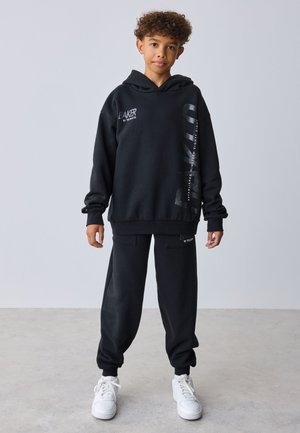 Enfant aux cheveux bouclés portant un sweat à capuche noir oversize et un jogging assorti avec des baskets blanches sur fond uni.