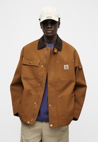 Άνδρας με καφέ εργασιακό μπουφάν με πολλές τσέπες, μπεζ παντελόνι cargo, λευκό καπέλο με το λογότυπο LA, σκούρα γυαλιά ηλίου και μπλε πουκάμισο από κάτω.