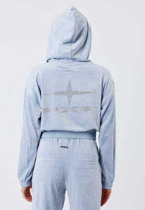 Mono con capucha azul claro de textura aterciopelada. Presenta un diseño de rhinestones en forma de estrella en la parte posterior y puños elásticos en las muñecas y la cintura.