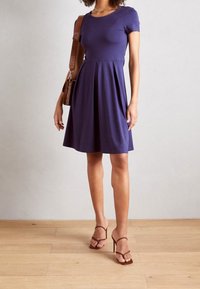 Abito blu navy a maniche corte con corpino aderente e gonna svasata. Tessuto liscio, lunghezza al ginocchio, abbinato a tacchi a spillo marroni e a una borsa marrone.