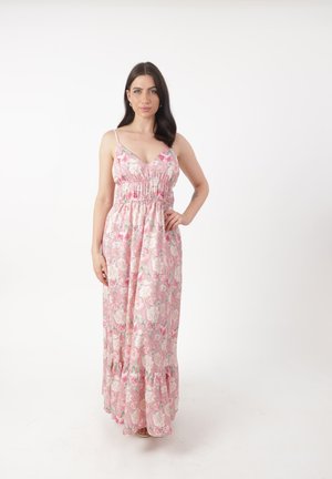 Vrouw met donker haar, gekleed in een roze maxijurk met bloemenprint zonder mouwen, staand met één hand op de heup tegen een effen witte achtergrond.