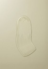 Texture gélatineuse claire, légèrement courbée, avec de douces nervures parallèles, placée sur un fond beige clair, ressemblant à un produit liquide.