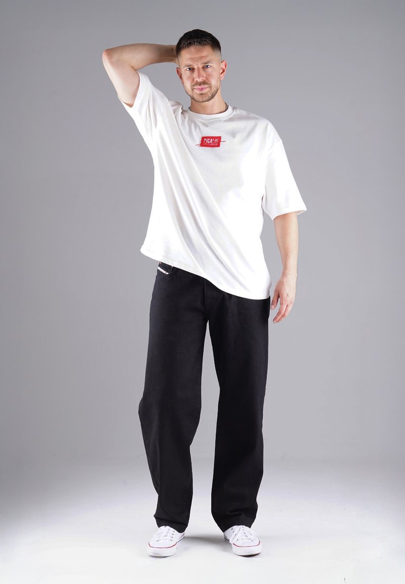 Weißes T-Shirt mit kurzen Ärmeln und einem roten Logo-Patch, kombiniert mit schwarzen weit geschnittenen Hosen und weißen Sneakern, vor einem grauen Hintergrund.