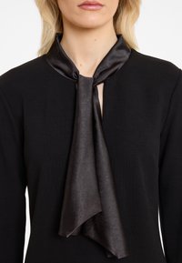 Schwarzes Oberteil mit einer Satin-Krawatte. Die Krawatte hängt mit einer glatten Textur herab. Der Ausschnitt ist mit einem Schlüssel-Design für zusätzliches Detail versehen.