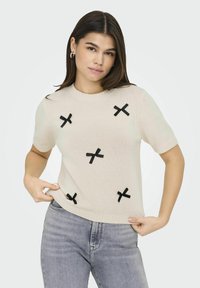 Brede gebreide trui in beige met korte mouwen, voorzien van zwarte strikdetails die over de voorkant zijn gerangschikt. Ronde halslijn en een relaxte pasvorm.