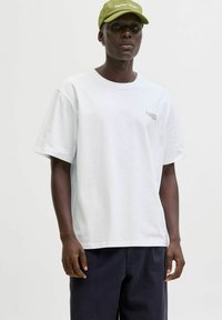 Hvidt bomuld t-shirt med korte ærmer, forsynet med et lille rødt logo på brystet. Båret sammen med navyblå løstsiddende bukser og en grøn kasket.