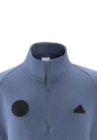 Pullover mezza zip in tessuto azzurro chiaro, con colletto alto, toppa rotonda nera e logo Adidas nero sul petto. Tessuto liscio.