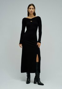 Schwarzes, langärmliges, figurbetontes Kleid mit Rundhalsausschnitt und Ausschnitt an der Brust, ausgestattet mit einem Seitenschlitz und gerippter Textur. Trägt schwarze Stiefel.