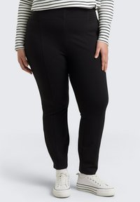 Zwarte stretchbroek met een gladde textuur en hoge tailleband. Gecombineerd met witte sneakers met zwarte strepen.