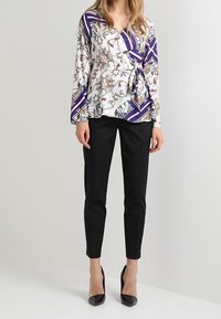 Top avvolgente multicolore con motivi floreali e geometrici, dotato di maniche lunghe e vita legata. Abbinato a pantaloni neri slim e tacchi.