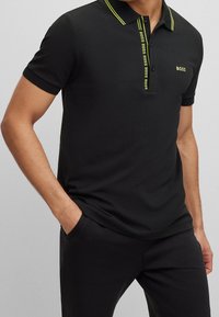 Hombre vistiendo una camiseta polo negra de manga corta con el logo y ribete amarillo de "BOSS", combinado con pantalones negros, con una mano en el bolsillo, de pie contra un fondo gris.