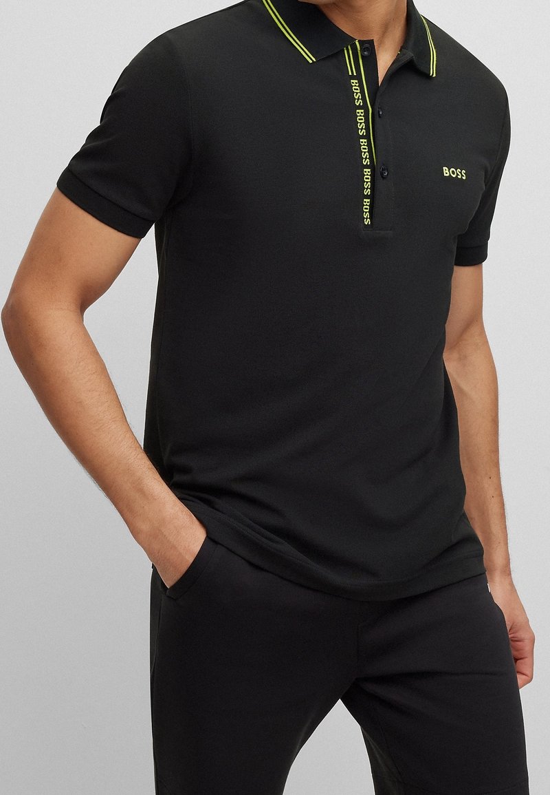 Hombre vistiendo una camiseta polo negra de manga corta con el logo y ribete amarillo de "BOSS", combinado con pantalones negros, con una mano en el bolsillo, de pie contra un fondo gris.