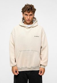 Beige hoodie met een voorzak, zachte textuur en een klein geborduurd logo op de borst. Losse pasvorm en verstelbare capuchon.