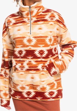 Personne portant un pull polaire chaud avec col montant, demi-fermeture éclair, poche avant, et motif géométrique orange, crème et rouge.
