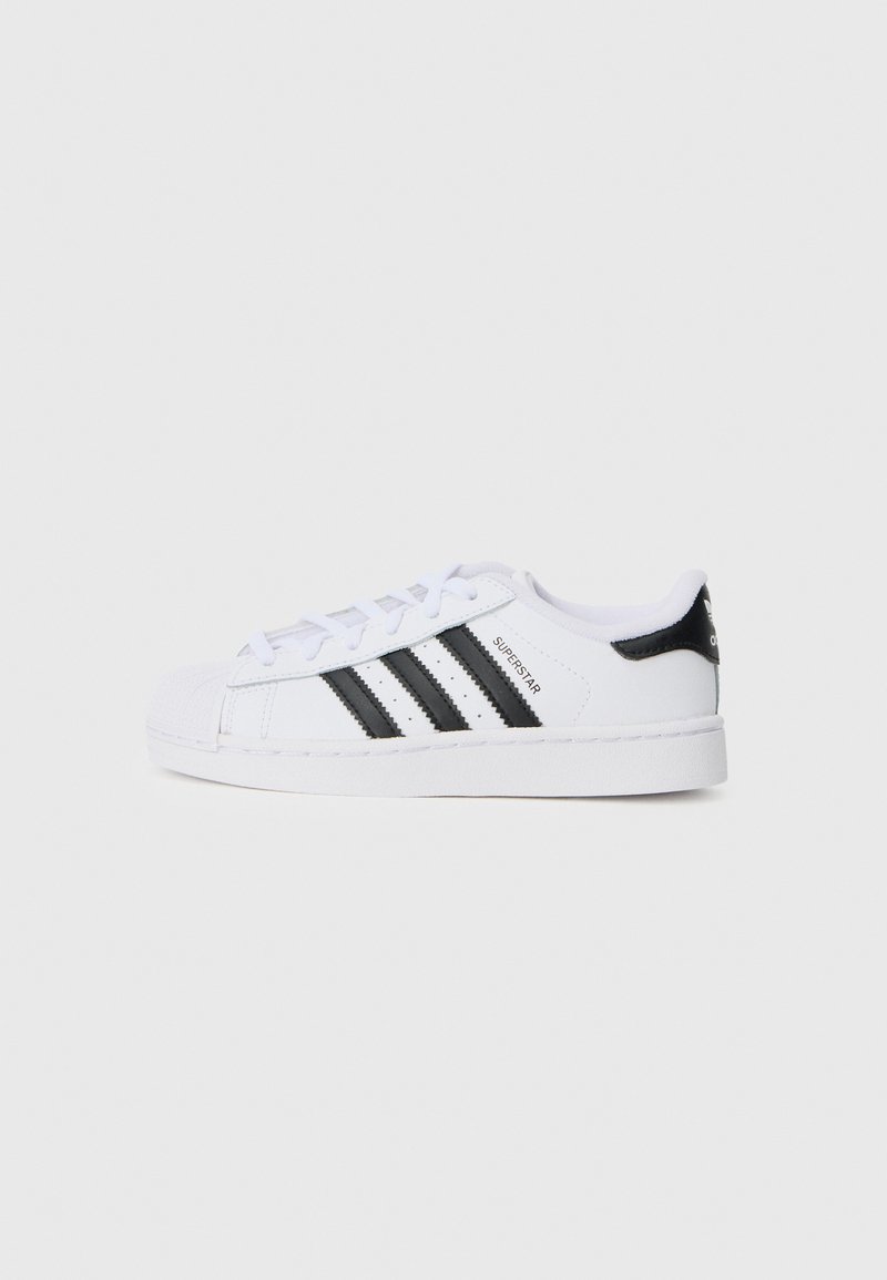 Άσπρα παπούτσια Adidas Superstar διαθέτουν τρεις μαύρες ρίγες, καπάκι από καουτσούκ στη μύτη και ανάγλυφη σόλα με λείο δερμάτινο επάνω μέρος.