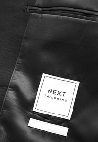 Intérieur de veste de costume noire avec une étiquette rectangulaire blanche portant la mention "NEXT TAILORING" cousue à l'intérieur.