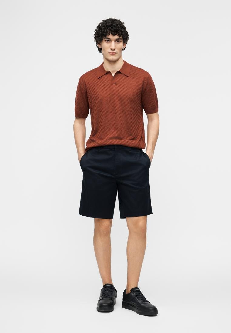 Man standing with hands in pockets wearing a rust short-sleeve polo, black shorts, and black sneakers on a plain background.