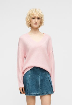 Νεαρή γυναίκα με ξανθά σγουρά μαλλιά που φοράει ροζ πουλόβερ με V λαιμόκοψη και μπλε τζιν πλισέ φούστα, στέκεται μπροστά σε απλό φόντο.