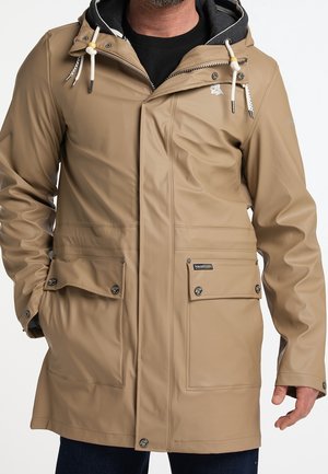 Mann trägt eine beige wasserdichte Jacke mit großen Vordertaschen, Kordelzugkapuze und schwarzem Futter, steht mit den Händen in den Taschen.