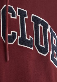 Vinröd sweatshirt med stor marinblå broderad text "CLUB" med vit kant. Mjuk textur, kontrasterande färger och hållbar konstruktion.