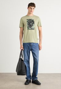 Lys grøn t-skjorte i bomull med grafisk trykk, kombinert med blå denimjeans og svarte joggesko. En svart tote bag holdes i den ene hånden.