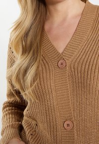Brązowy, prążkowany kardigan dzianinowy z dekoltem w serek, długimi rękawami i dwoma dużymi, zaokrąglonymi guzikami z przodu. Miękka tekstura, swobodny design.