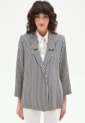 Femme aux cheveux bouclés et foncés portant un blazer à rayures verticales noires et blanches sur une chemise blanche, debout devant un fond uni.