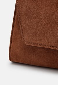 Brun ruskinds håndtaske med en trekantet klap, pæne syninger, blød tekstur og en struktureret form. Nærbillede der fremhæver materialedetaljer.