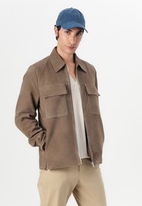 Brun ruskindsjakke med to frontlommer, lynlås og spids krave, båret over en lys grå sweater og beige bukser. En blå kasket fuldender looket.