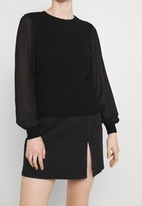 Svart långärmad topp med genomskinliga ärmar och ribbade manchetter, tillsammans med en svart denim minikjol med en sidoklyfta.