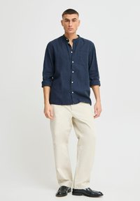 Camisa de lino azul marino con cuello abotonado, bolsillo delantero y cierre de botones, combinada con pantalones crema y zapatos negros.