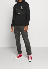 Czarna bluza z kapturem z zieloną grafiką, czarna nerka, szare dżinsy oraz czerwone trampki za kostkę z białą podeszwą.