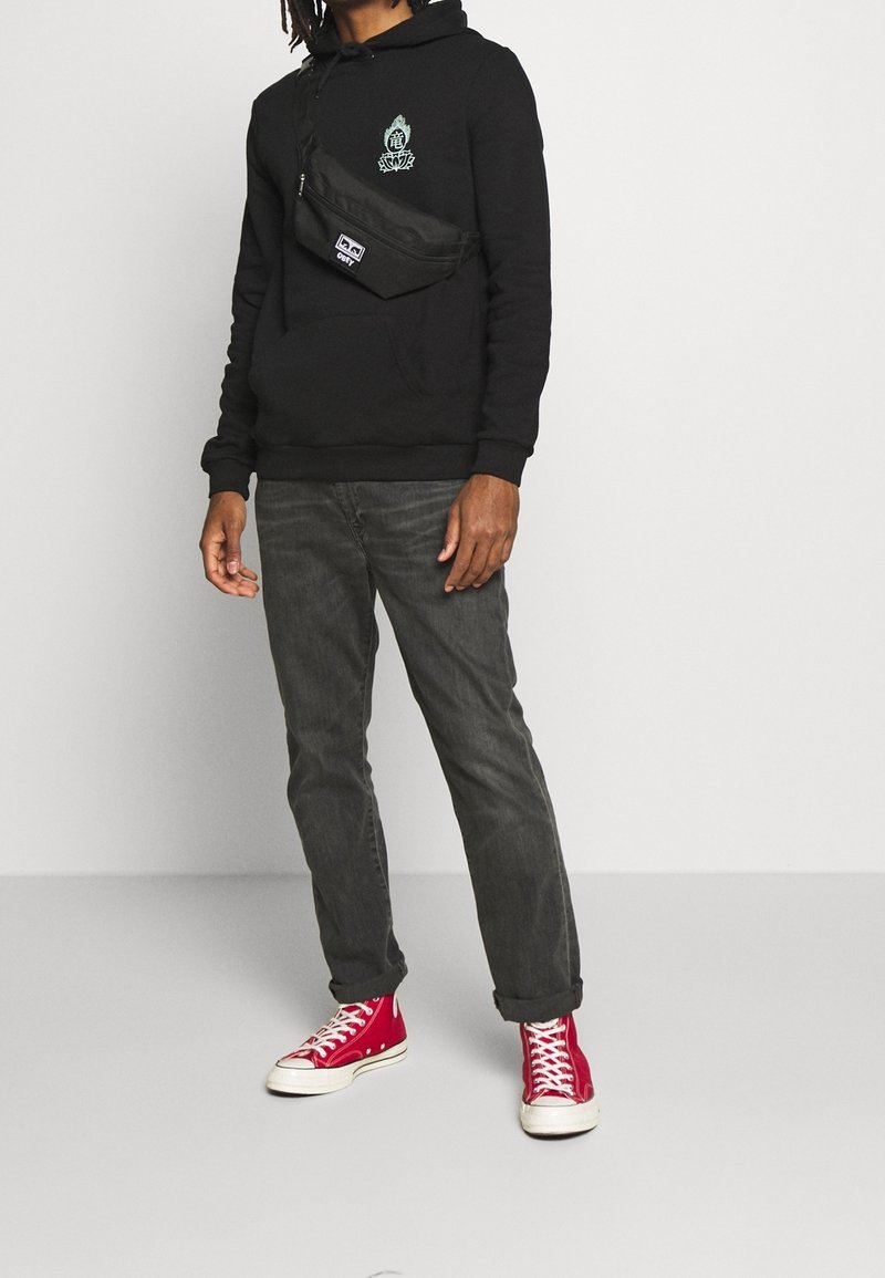 Czarna bluza z kapturem z zieloną grafiką, czarna nerka, szare dżinsy oraz czerwone trampki za kostkę z białą podeszwą.