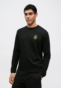 Czarna koszula z długim rękawem wykonana z miękkiego materiału, z pozłacanym logo na piersi. Prosty krój z okrągłym dekoltem i dopasowanymi rękawami.