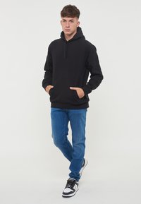 Felpa nera con tasche a marsupio e cappuccio regolabile, abbinata a jeans blu e sneakers bianco e nere, in piedi contro uno sfondo bianco.