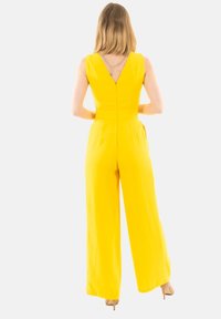 Tuta gialla con schiena a V, gambe larghe e dettagli in catena argentata sul colletto. Realizzato in un tessuto morbido con vita aderente.