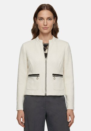 Femme portant une veste blanche texturée zippée avec des poches zippées noires, debout devant un fond clair uni.