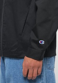 Svart jakke med en glatt tekstur, med en logo på mansjetten, kombinert med lyseblå denimbukser. Glidelåsdetalj delvis synlig.