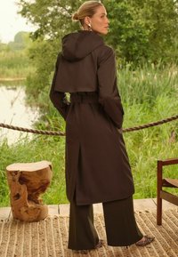 Rjava vodoodporna jakna s kapuco, z pasom in razširjenim robom, v kombinaciji z širokimi hlačami in čevlji z živalskim vzorcem, na naravni podlagi.