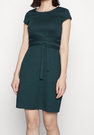 Kobieta w krótkiej sukience z ciemnozielonego materiału z przewiązanym paskiem w talii, stojąca na tle jednolitego białego tła.