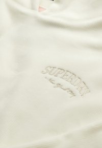 Tela de color crema con un sutil bordado del logo "SUPERDRY" y un texto en cursiva más pequeño debajo, todo sobre un material suave y texturizado.